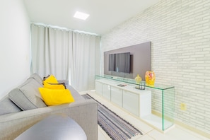 A modern living room with a grey sofa, yellow cushions, a glass coffee table, a flat-screen TV on a white brick wall, and a glass cabinet with decorative items. - Porto Plaza #210 - Triple Flat by Carpediem (Ipojuca)