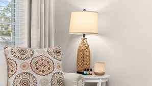 Ein Zimmer mit einem gemusterten Kissen, einer Lampe mit geflochtenem Fuß und einem kleinen Tisch mit Kerze und ätherischen Ölen.
