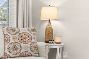 Ein Zimmer mit einem gemusterten Kissen, einer Lampe mit geflochtenem Fuß und einem kleinen Tisch mit Kerze und ätherischen Ölen.