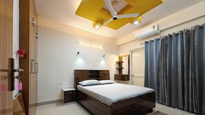 A modern bedroom with a wooden bed, a ceiling fan, a bedside table, and a large window with curtains. - Casa InteliStay – Smart, Stylish 2BHK in Lucknow (Lucknow)