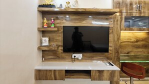 A modern living room with a large flat-screen TV mounted on a wooden wall, a small shelf with decorative items, and a wooden cabinet with a TV on top. - Casa InteliStay – Smart, Stylish 2BHK in Lucknow (Lucknow)