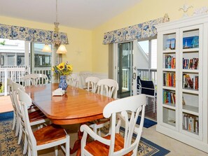 A dining area with a wooden table and white chairs, a bookshelf with books, and a view through a glass door to the outside. - 4BR House w/ Deck | Close to Beach! (South Bethany Beach)