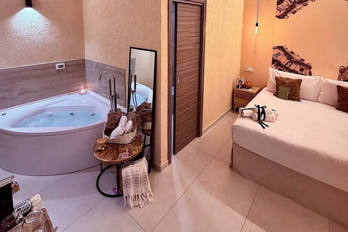 A modern bathroom with a freestanding bathtub, a vanity with a mirror, and a bedside table with a lamp.
