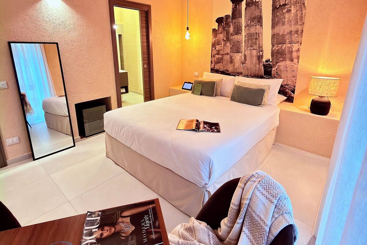 A hotel room with a large bed, a bedside table with a magazine, a lamp, and a wall-mounted TV.