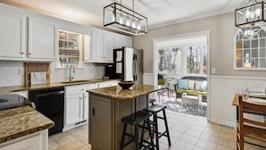 A kitchen with white cabinets, a granite countertop, a central island with stools, a refrigerator, and a dining area with a table and chairs. - 3BR House near Bethany Beach | Pool Access (Frankford)