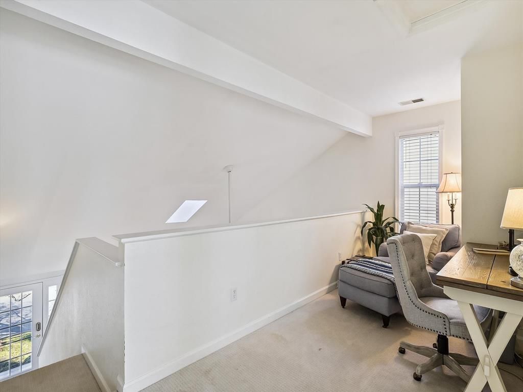 Une pièce spacieuse avec un plafond haut, un escalier, un bureau avec une lampe et une fenêtre équipée de stores.
