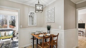 A dining area with a wooden table set for four, white paneled walls, a decorative wall piece, and a window with a view of trees. - 3BR House near Bethany Beach | Pool Access (Frankford)