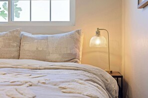Ein Schlafzimmer mit einem Bett, Kissen, einem Nachttisch, einer Lampe und einem Fenster mit Blick auf Bäume.