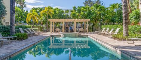 A swimming pool with lounge chairs, a pergola, and palm trees.