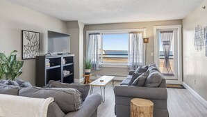 Una sala moderna con un sofá gris, un televisor de pantalla plana sobre un mueble bajo, una mesita de madera y vistas a la playa a través de grandes ventanales.