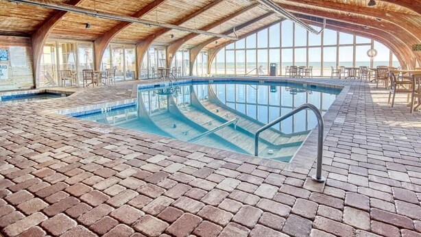 An indoor swimming pool with a brick floor and wooden ceiling, surrounded by tables and chairs. - Carol's Coastal Getaway (Myrtle Beach)