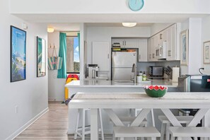 A modern kitchen with white cabinets, a stainless steel refrigerator, and a microwave. There is a dining table with benches and a green bowl on it. - Sands Beach Club 2BR Ocean Front Condo 728 (Myrtle Beach)