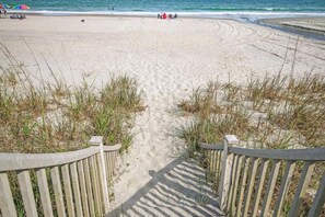 Une plage de sable avec une promenade en bois menant à l’eau, entourée d’herbe et bordée d’une rambarde.