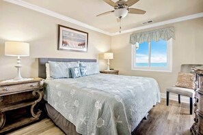 Une chambre à coucher comprenant un lit, deux tables de chevet, un ventilateur de plafond, une fenêtre avec vue et un coussin décoratif orné d’un motif.