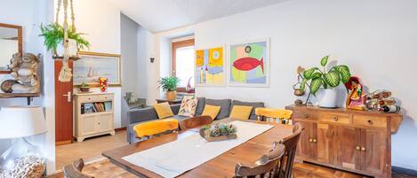 Una sala acogedora con una mesa de madera para comer, asientos cómodos, un sofá con cojines y una estantería.