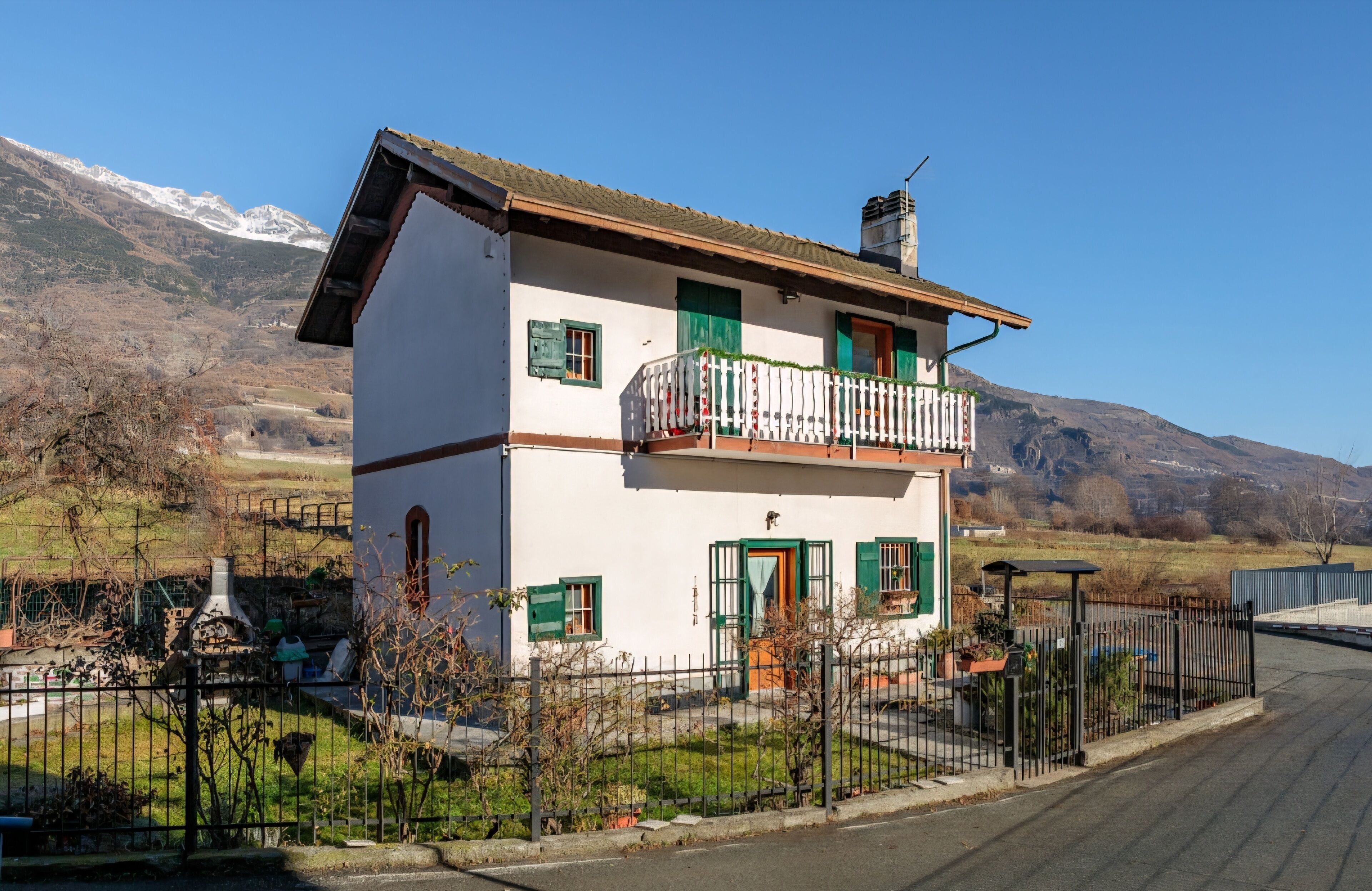 Ein zweistöckiges Haus mit Balkon, umgeben von einem Zaun, vor einer Bergkulisse.