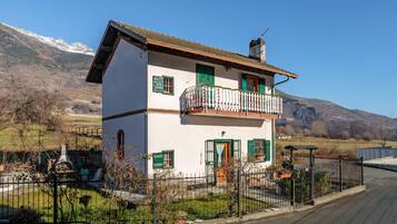 Ein zweistöckiges Haus mit Balkon, umgeben von einem Zaun, vor einer Bergkulisse.