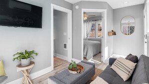 A modern living room with a grey sofa, a small side table with a plant, and a flat-screen TV mounted on the wall. - Bright and Tranquil Guesthouse! (Woodfin)