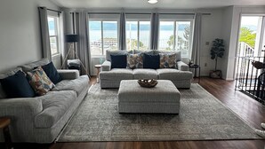 Ein geräumiges Wohnzimmer mit einer grauen Sofagarnitur, einem Couchtisch und einem Deckenventilator. Große Fenster bieten Blick auf ein Gewässer und Berge.
