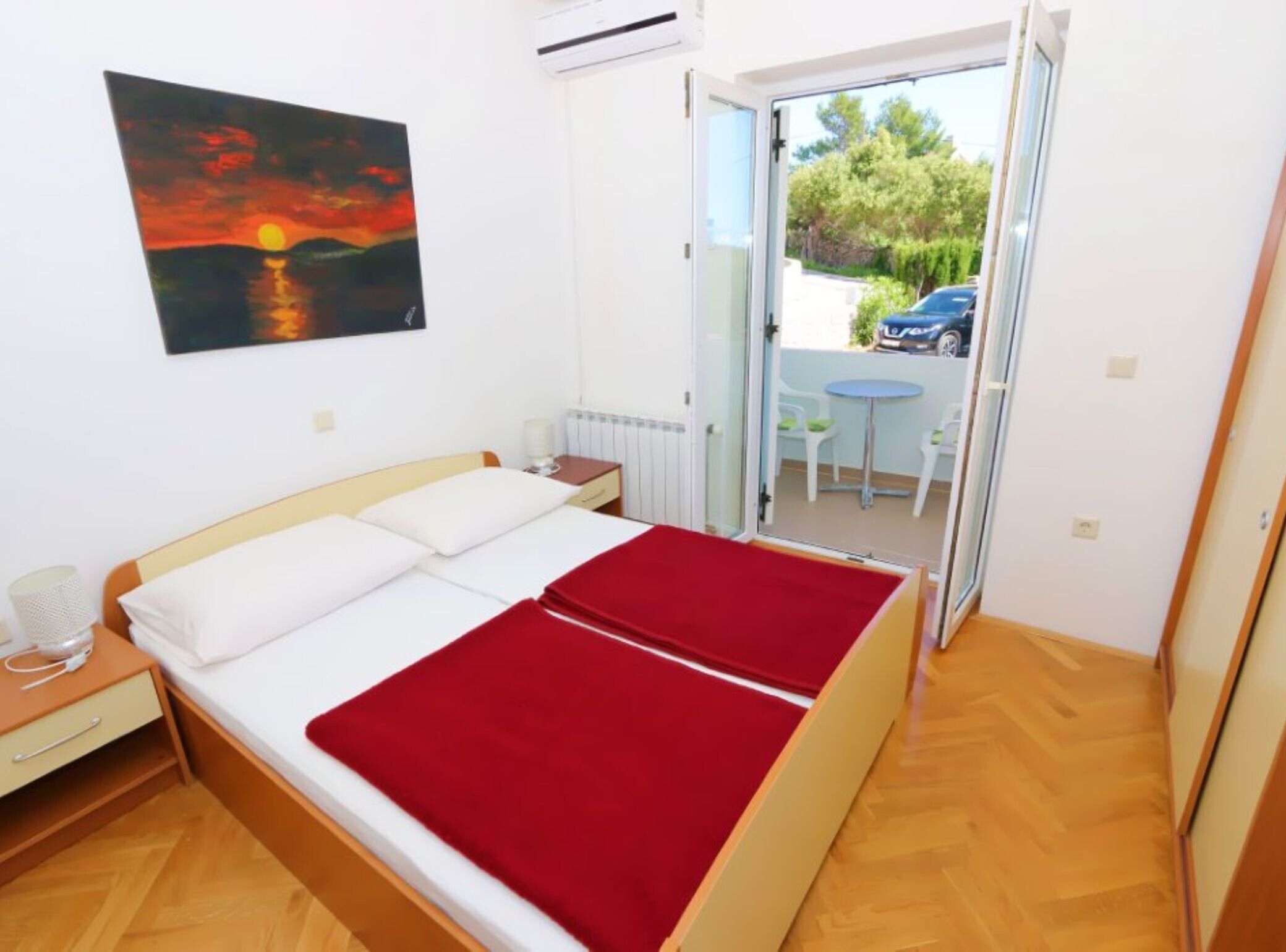 A bedroom with a bed, a nightstand, a red blanket, a painting of a sunset, and a view of a car and greenery outside.