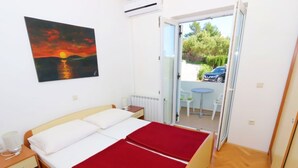 Une chambre avec un lit, une table de chevet, une couverture rouge, un tableau représentant un coucher de soleil et une vue sur une voiture et de la verdure à l’extérieur.