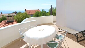Un balcon avec une table ronde et des chaises, donnant sur une zone côtière avec des maisons et un ciel dégagé.