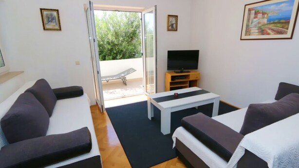 Quarto moderno com sofá, aparador para TV e uma pequena mesa. Há uma porta que dá para área externa com um banco.