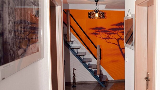 Una escalera con barandilla de madera y peldaños de metal, que lleva a una habitación con pared naranja y un mural de un árbol.