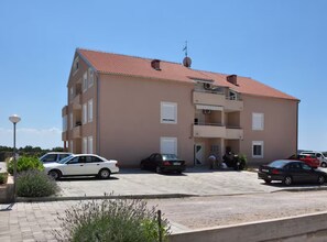 A multi-story building with a red-tiled roof, a parking area with several cars, and a clear blue sky. - Apartments Ad Astra - Two Bedroom Apartment with Terrace and Sea View (Vodice)