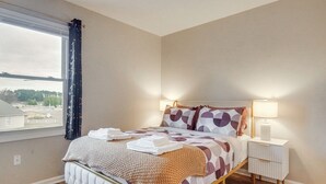 A neatly made bed with a patterned bedspread and pillows, a bedside table with a lamp, and a window with curtains. - 3BD Townhome w/ Patio, 3 Mi to Fort Bragg/Liberty (Fayetteville)