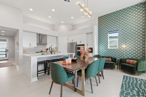 A modern kitchen with white cabinets, a central island, and a dining area with green chairs. - Casa Volare- Pool+Spa, Gated 4BR 3BA 2,400sqft Brand New Desert Retreat (Bermuda Dunes)