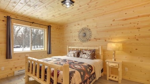 A wooden cabin bedroom with a bed, a window, a decorative wall piece, and a nightstand with a lamp. - Retreat Near Ice Fishing, ATV Trails, Golf, Skiing (Danbury)