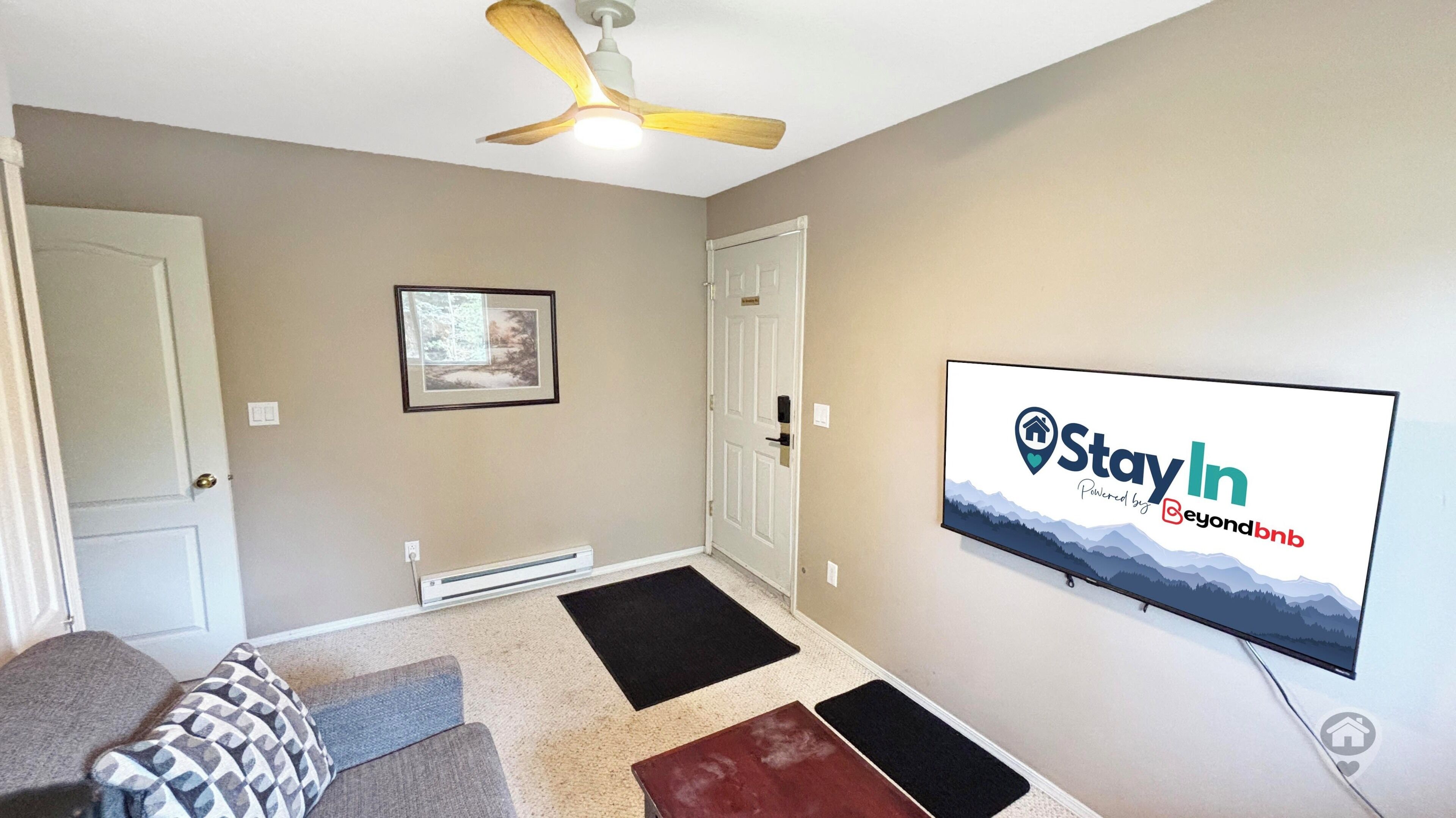 Una sala de estar con ventilador de techo, un televisor montado mostrando un anuncio de 'StayIn', un cuadro en la pared y una puerta.