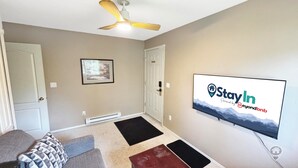 A living room with a ceiling fan, a mounted TV displaying a 'StayIn' advertisement, a framed picture on the wall, and a door. - 1BR Suite with River & Mountain Views in Fernie (Fernie)