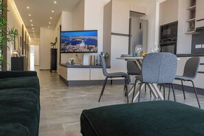 Un salon moderne avec un canapé, un coin repas comprenant une table et des chaises, un téléviseur à écran plat diffusant une vue sur la ville, et une cuisine équipée d’appareils électroménagers.