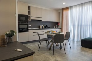 Una cocina moderna con electrodomésticos integrados, un comedor con mesa y sillas, y un amplio ventanal con cortinas translúcidas.