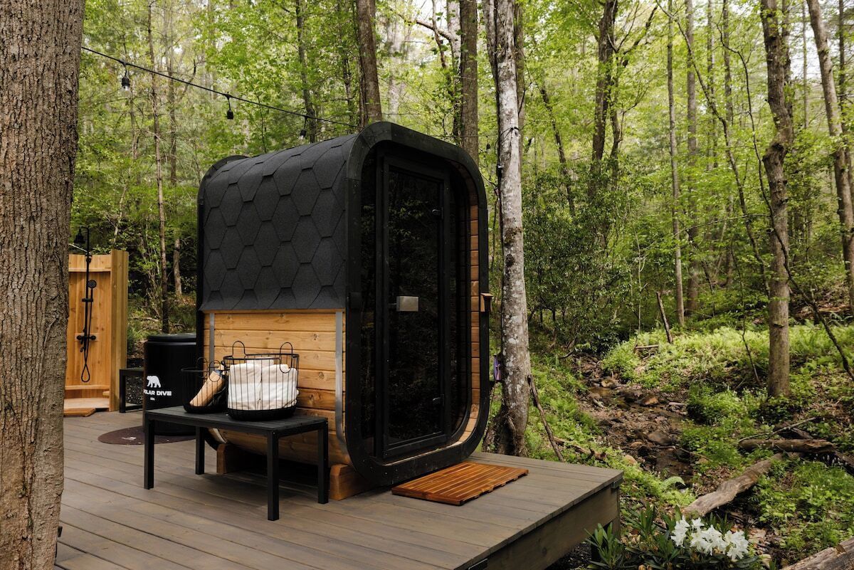 Une cabane en bois à six côtés, avec une façade noire, une terrasse en bois et un banc avec des sacs.