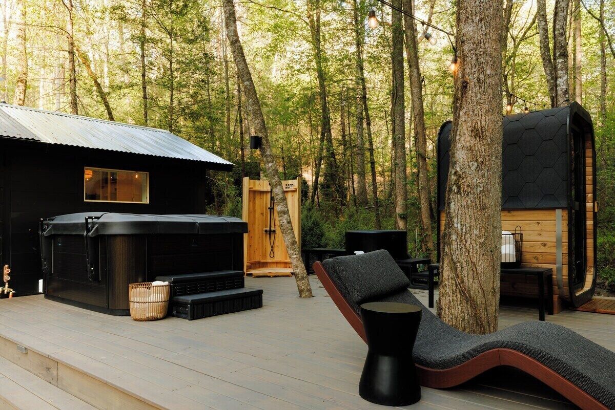 A wooden cabin with a hot tub, outdoor seating, and a deck surrounded by trees.