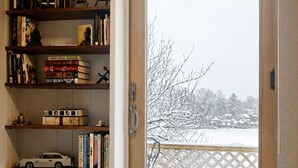 A cozy room with a large window overlooking a snowy landscape, a bookshelf filled with books and a model car, and a plush beige blanket on the bed.