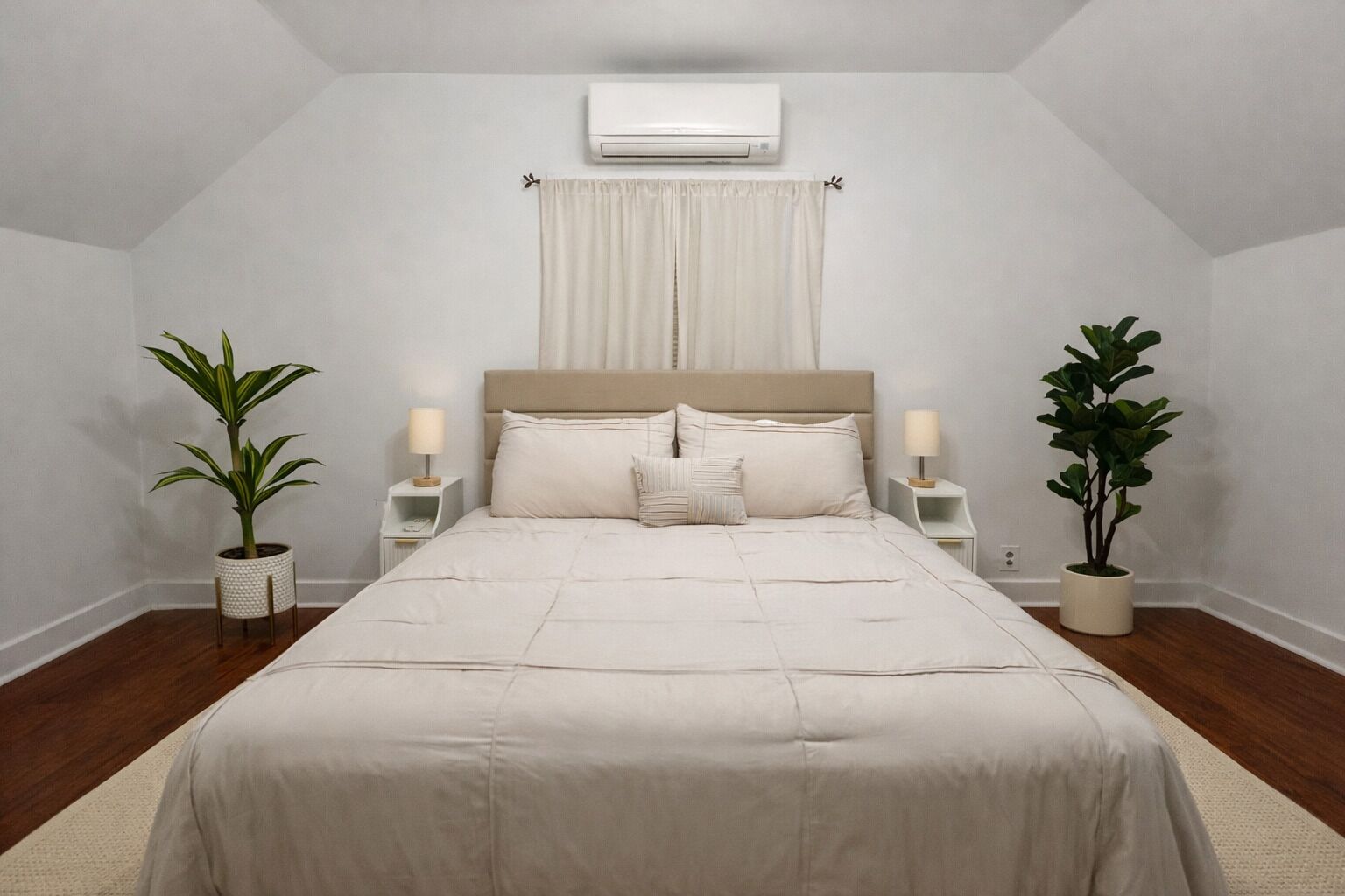 A bedroom with a bed, two bedside tables, a nightstand, a potted plant, a wall-mounted air conditioner, and a window with curtains.