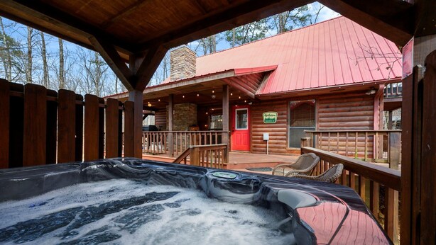Eine Holzhütte mit Whirlpool, einer Holzdar Terrasse und einem Gebäude mit rotem Dach im Hintergrund.