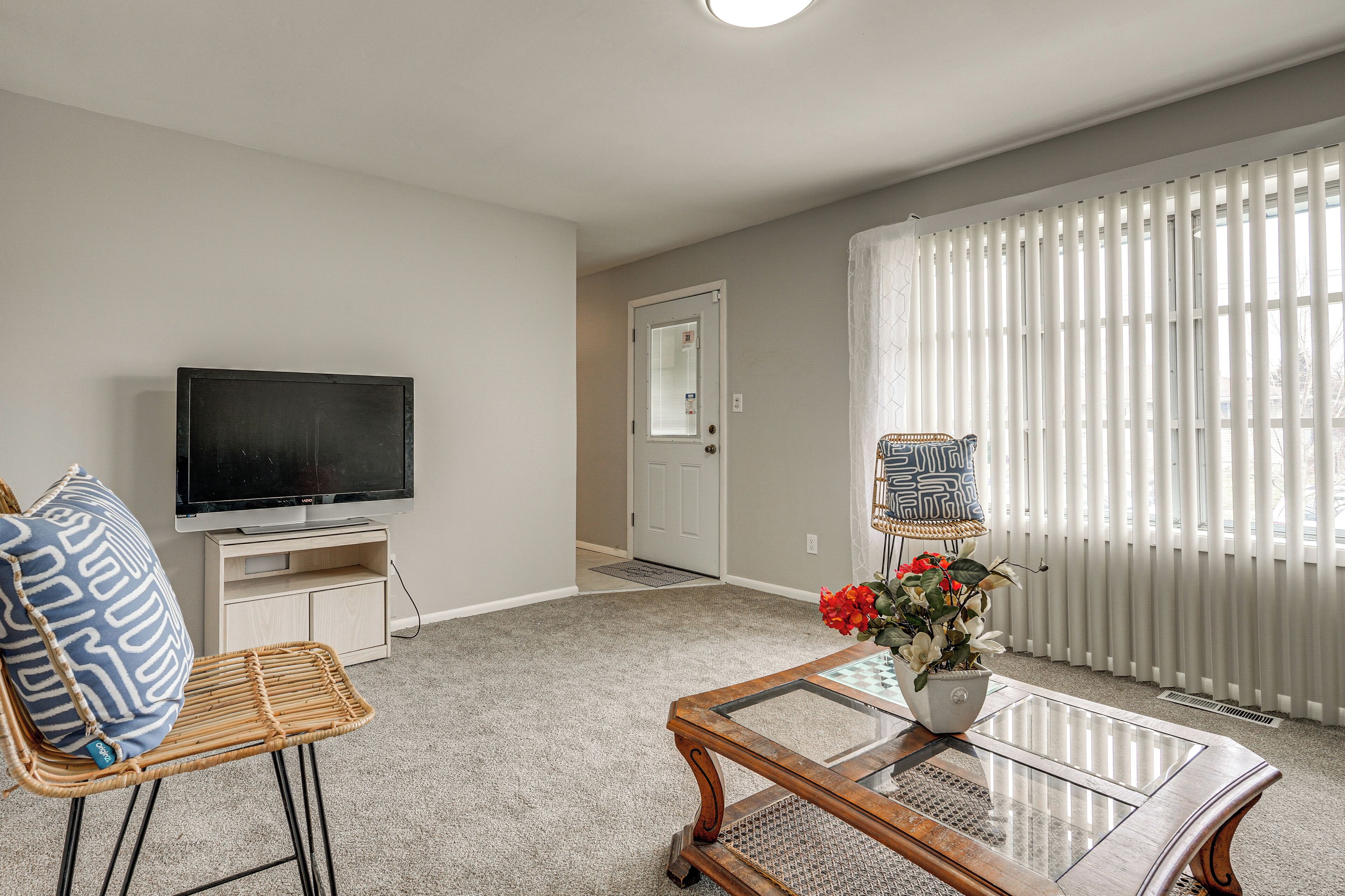 Una sala de estar con televisor, una silla de mimbre, una mesa de centro de vidrio y una puerta que da a otra habitación.