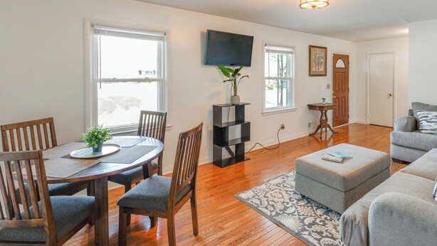 A living room with a dining table, chairs, a grey sofa, a TV mounted on the wall, and a wooden floor. - Near Medical Centers & Fort Jackson! Quiet Home (Columbia)