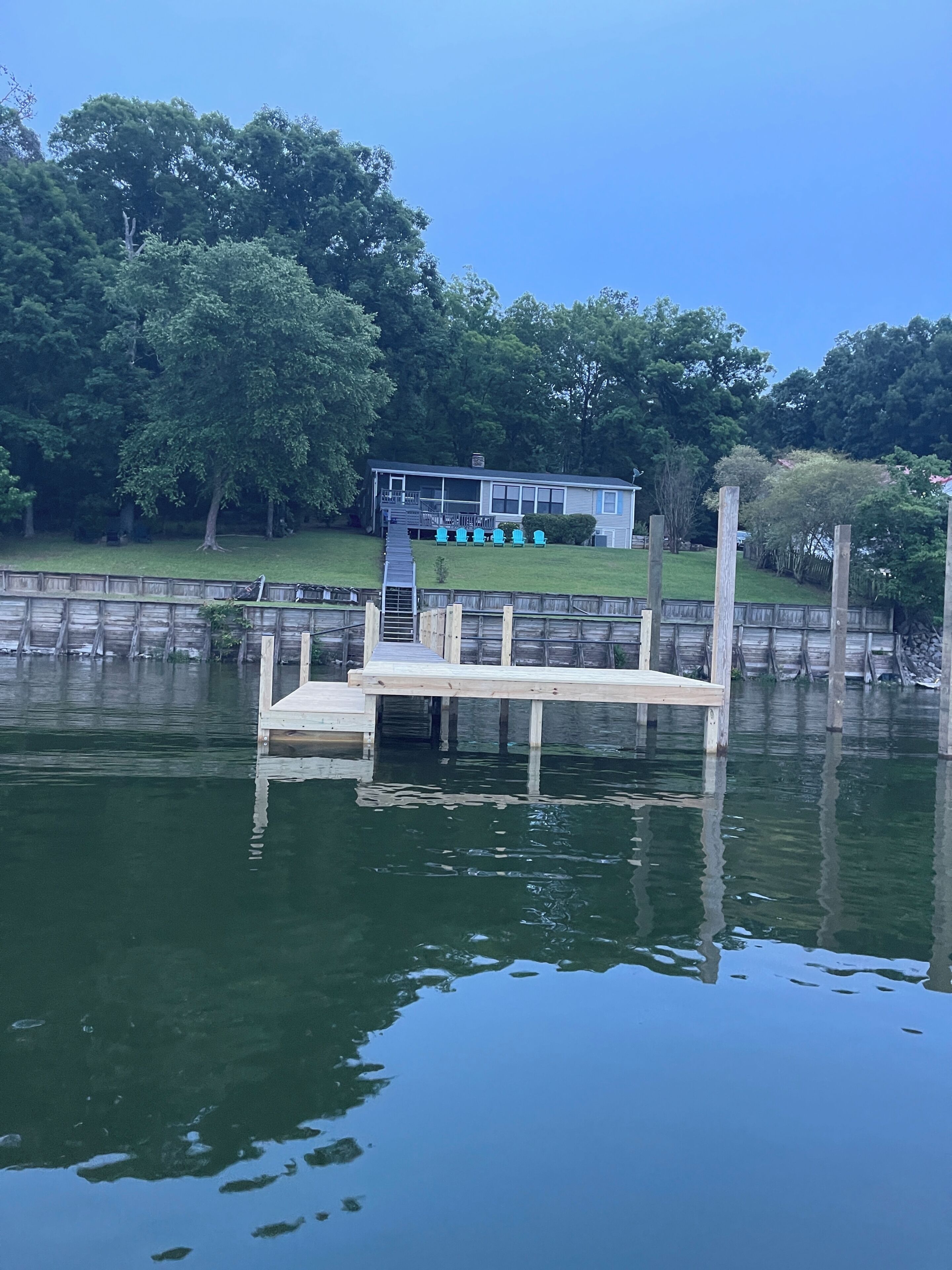 A wooden dock extending into a calm body of water, with a house and outdoor seating area in the background.