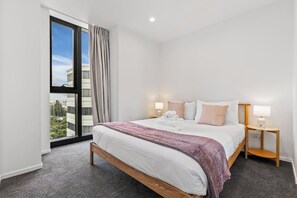 A hotel room with a large bed, two bedside tables with lamps, a view of the city through a large window, and a carpeted floor. - Modern High Rise w/ Parking by Zodiak Stays (Auckland)