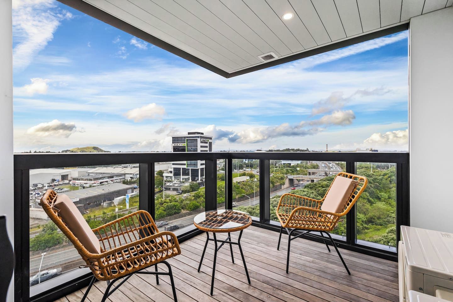 Een balkon met rieten stoelen en een tafeltje, met uitzicht op een stadsbeeld met gebouwen en groen.
