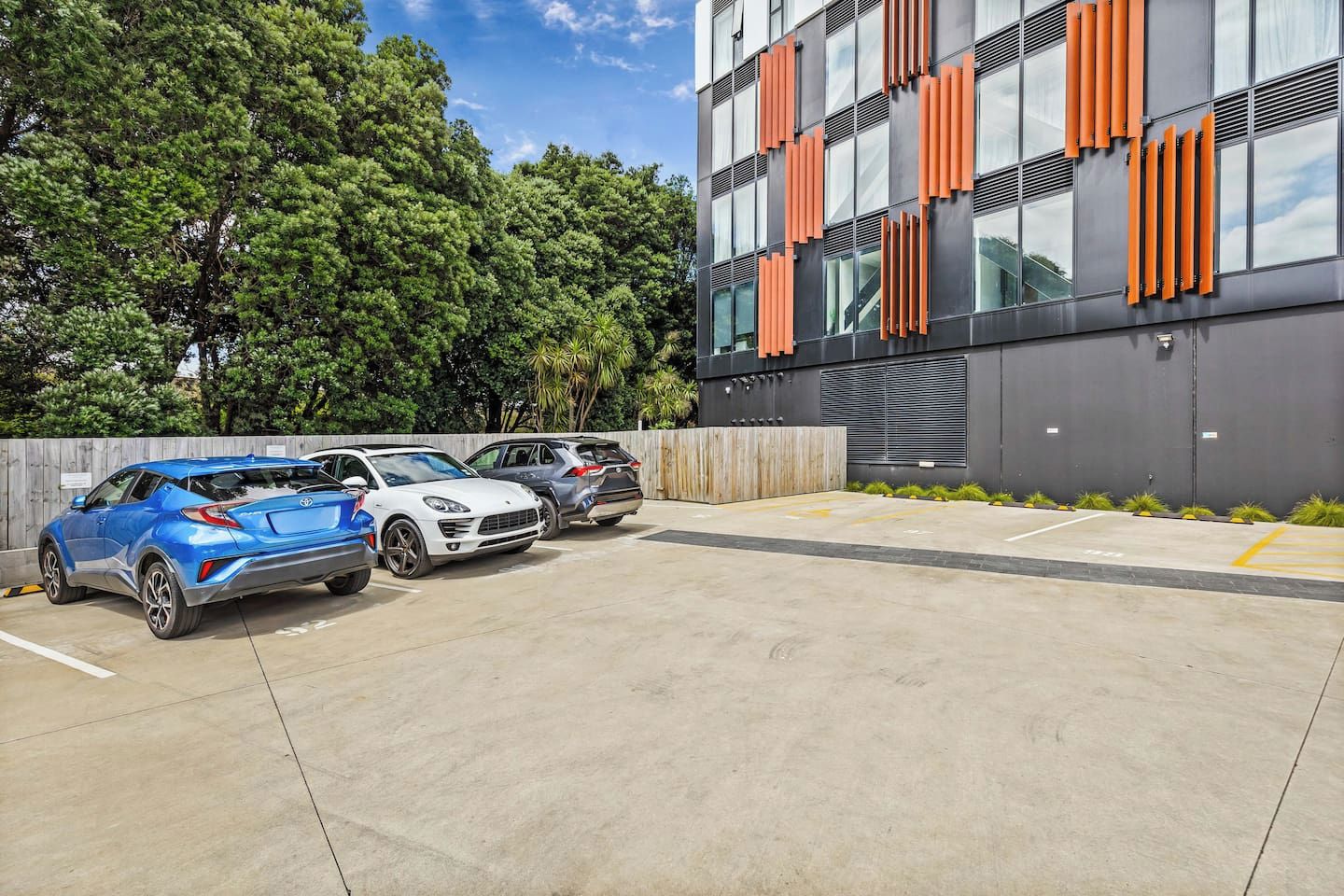 A parking area with three cars, a modern building with orange accents, and a backdrop of trees.