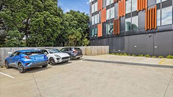 A parking area with three cars, a modern building with orange accents, and a backdrop of trees.