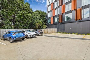 A parking area with three cars, a modern building with orange accents, and a backdrop of trees. - Modern High Rise w/ Parking by Zodiak Stays (Auckland)
