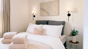 Una cama bien hecha con ropa blanca y una toalla doblada, una mesita de noche con lámpara, una obra de arte abstracta enmarco y una planta pequeña en maceta.